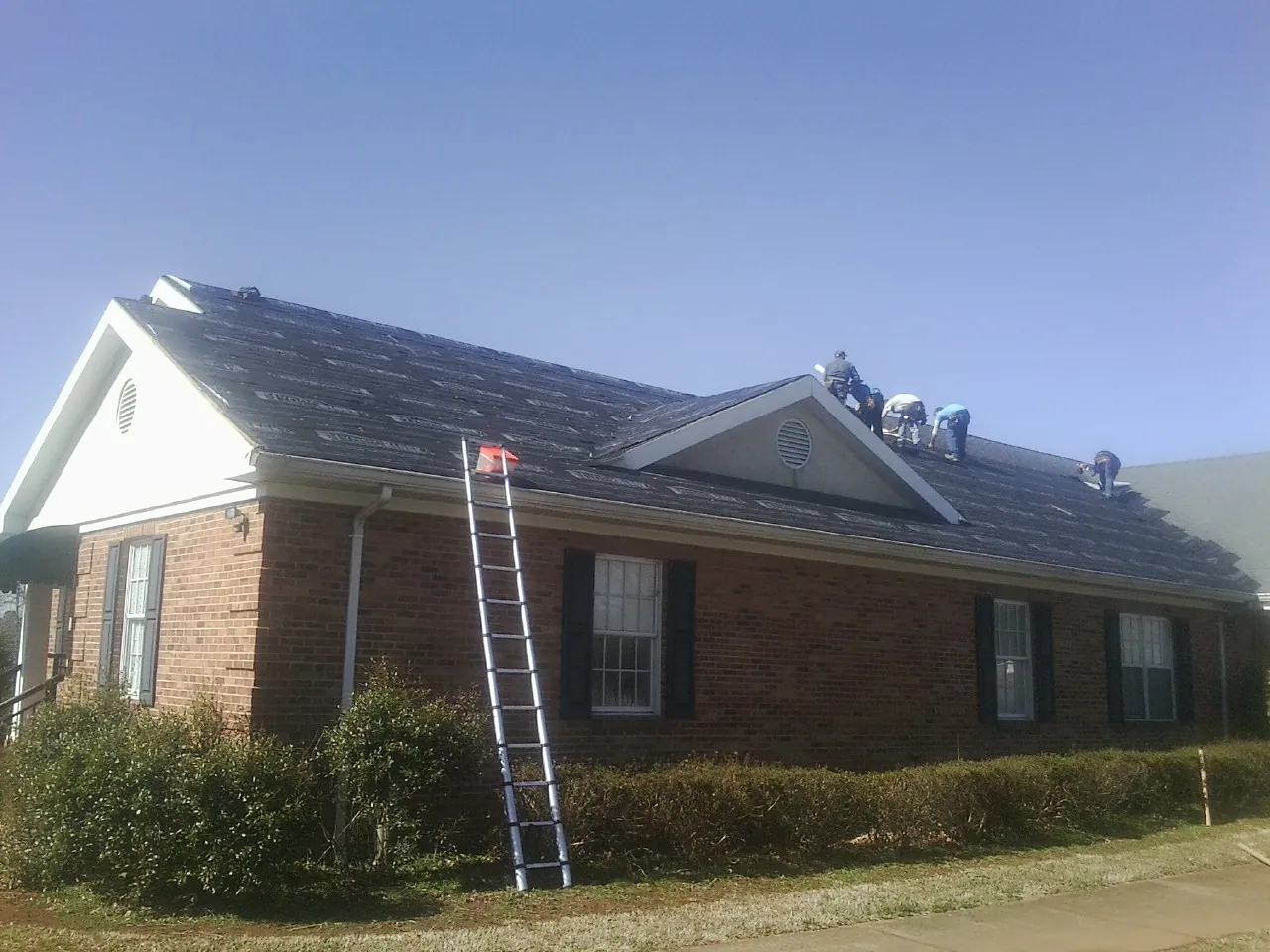 Crew tearing shingles off a brick ranch roof with a ladder set — Emergency Roof Repair work by our crew in Ireton