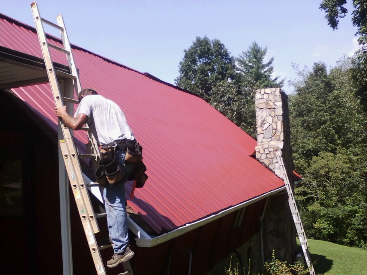 Roofer on a ladder beside a red metal roof and stone chimney — Soffit & Fascia Repair by Roofing Brothers in Ireton